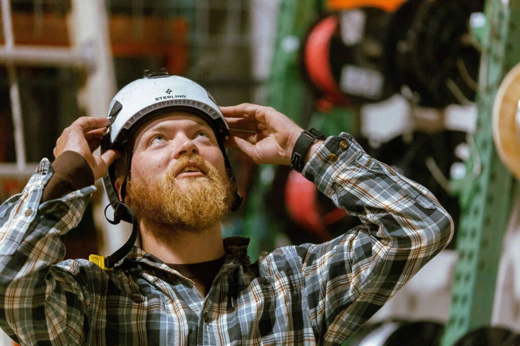 A bearded man in a plaid shirt adjusts a white safety helmet in an industrial workshop.